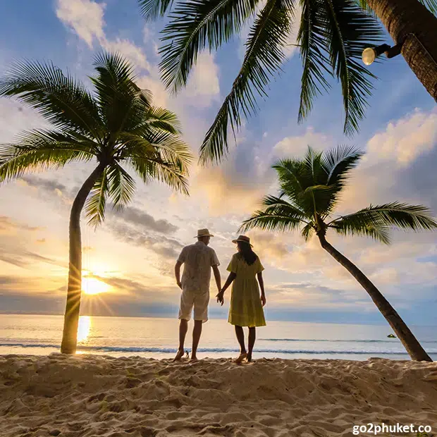 Couple sitting on Bang Tao Beach sand watching colorful sunset over the Andaman Sea in Phuket, Thailand