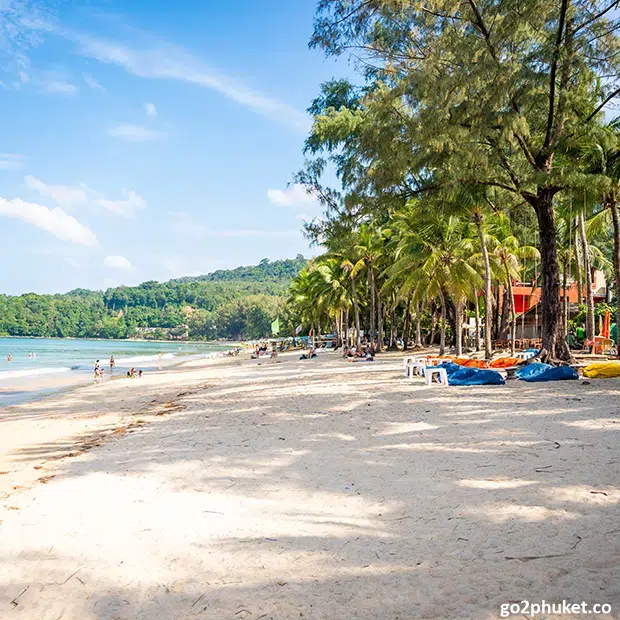 Visitors relaxing on deck chairs while bathers swim in calm turquoise water at Kamala Beach, Phuket Thailand.