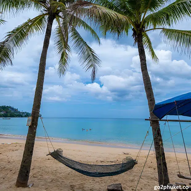 Colorful hammock tied between palm trees overlooking sandy Kamala Beach and calm Andaman Sea in Phuket Thailand.
