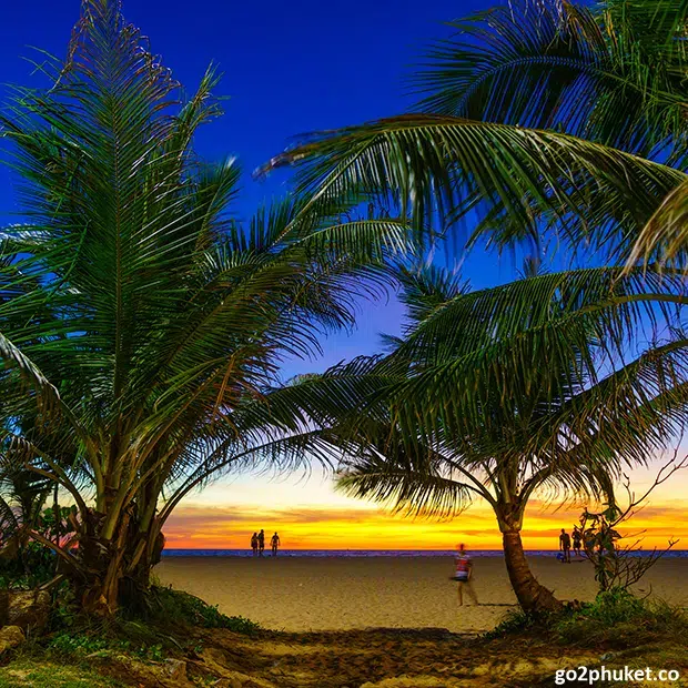 Visitors walking along Karon Beach shoreline during sunset with colorful sky over the Andaman Sea in Phuket Thailand.