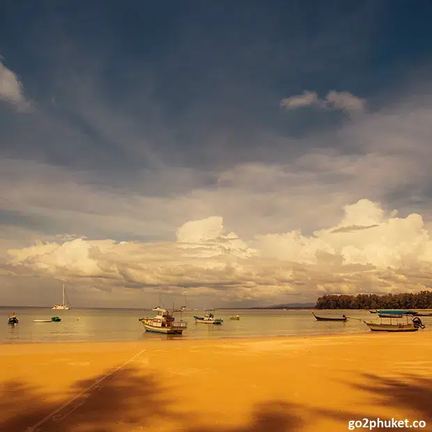 Calm shoreline at Nai Yang Beach with cloudy sky and fishing boats on the Andaman Sea in Phuket Thailand