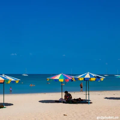 Bathers relaxing under umbrellas on Nai Yang Beach with a yacht anchored offshore in Phuket Thailand